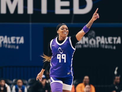 Odyssey Sims, wearing a purple #99 Athletes Unlimited basketball uniform, points to the sky in celebration and smiles as she runs up the court.