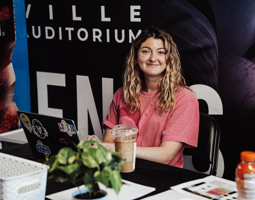AU Staff Member sitting at a table with computer and coffee in front. She is wearing a pink shirt and smiling at the camera