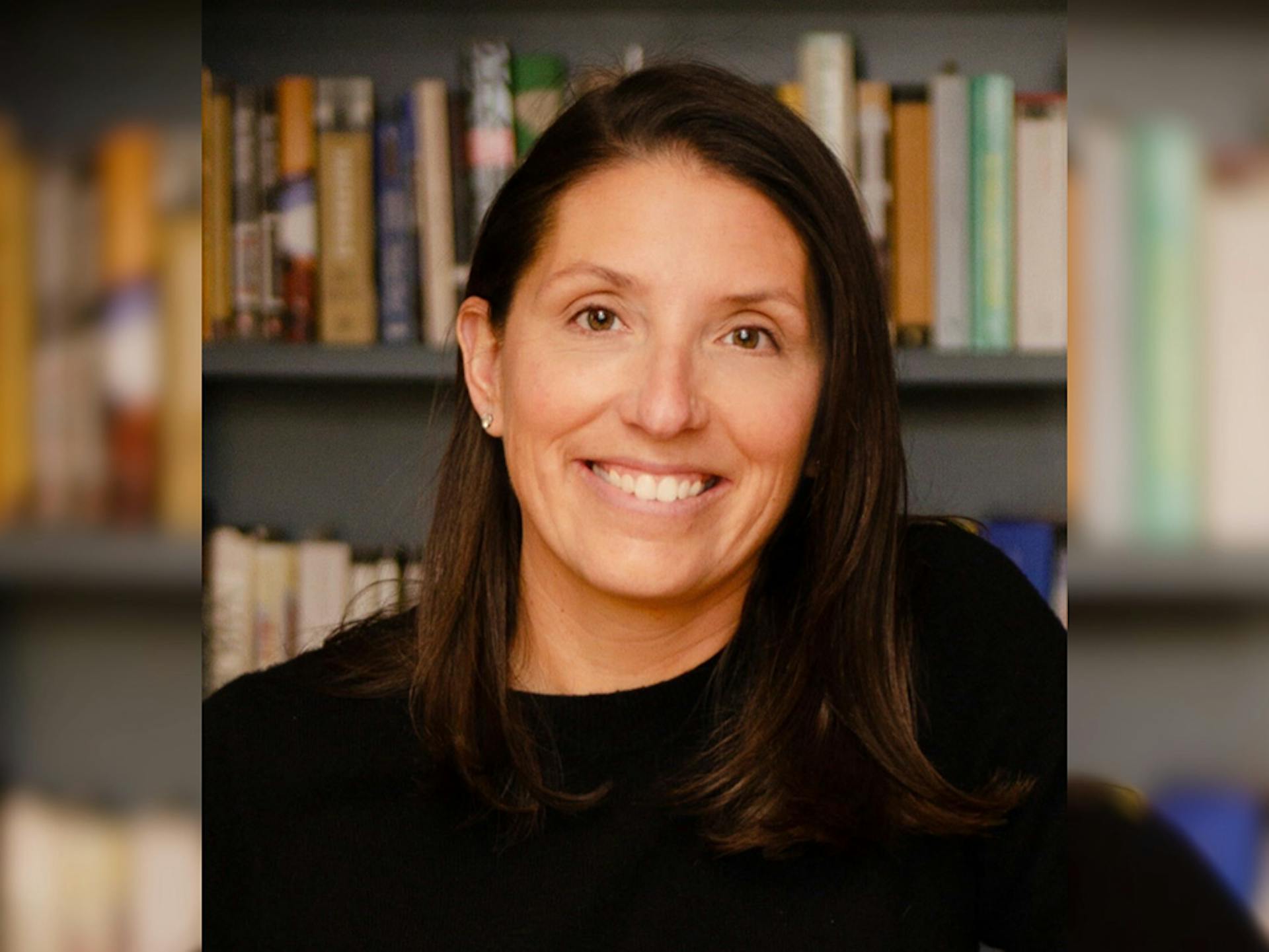 Allie Kleva, wearing a black top, smiles as she sits in front of a bookshelf.
