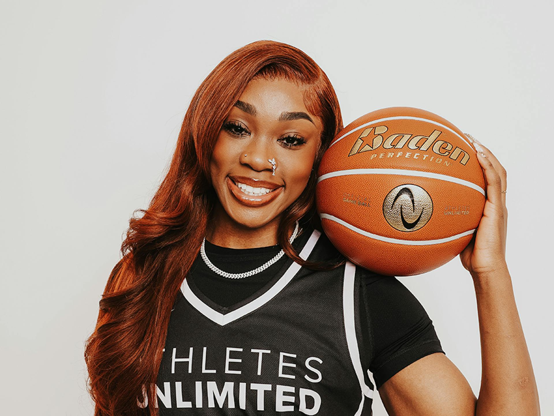 Aneesah Morrow, wearing a black AU Pro Basketball jersey with white trim and lettering, and a black short-sleeved undershirt, smiles as she rests a basketball on her shoulder with one hand.