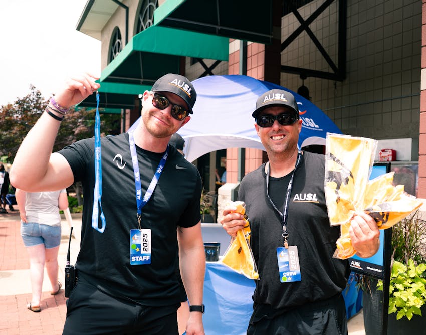 Two AUSL staff members smiling at the camera in black t-shirts, black hats and blue lanyards with AUSL