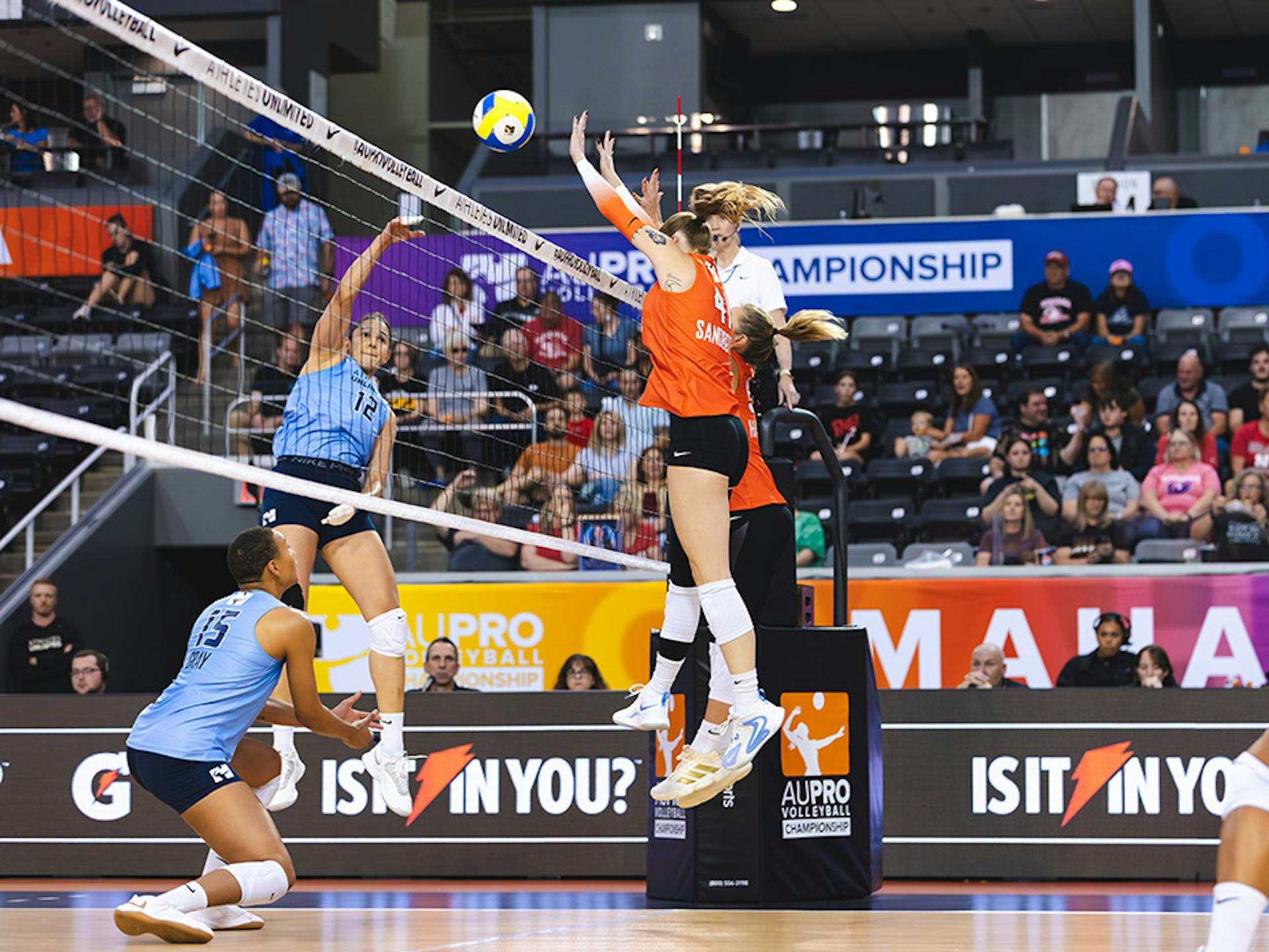 Natalia Zilio Pereira in a blue jersey hitting the ball over the net by two Team De La Cruz-Mejía players in orange jerseys